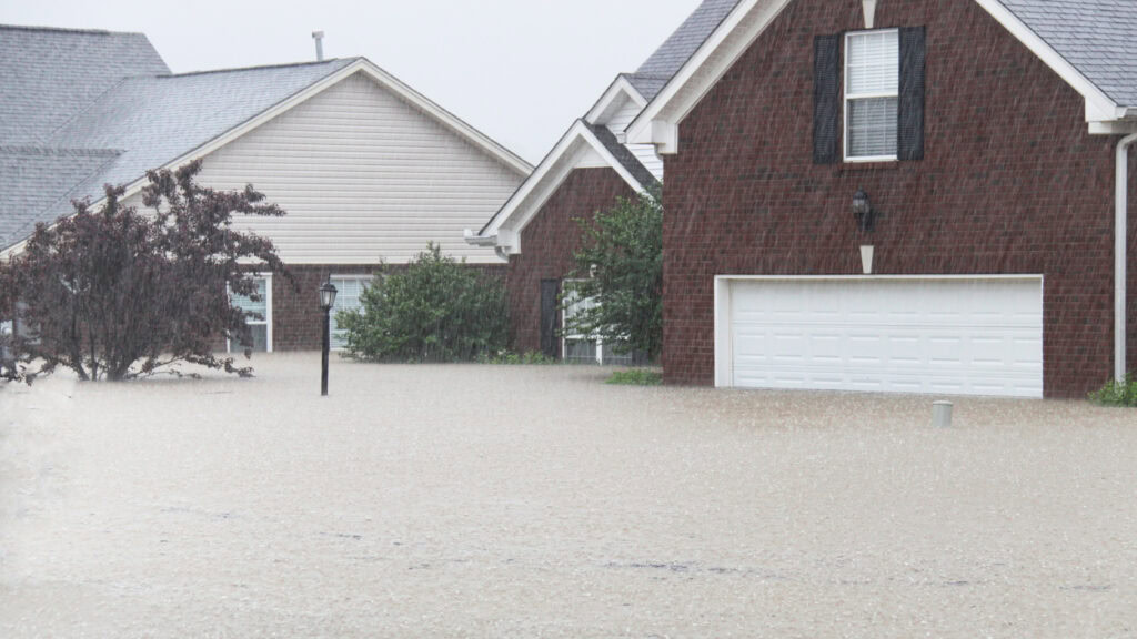 Flooded residential neighborhood with water covering the streets and part of the driveways. Two houses and some trees are visible under heavy rainfall.