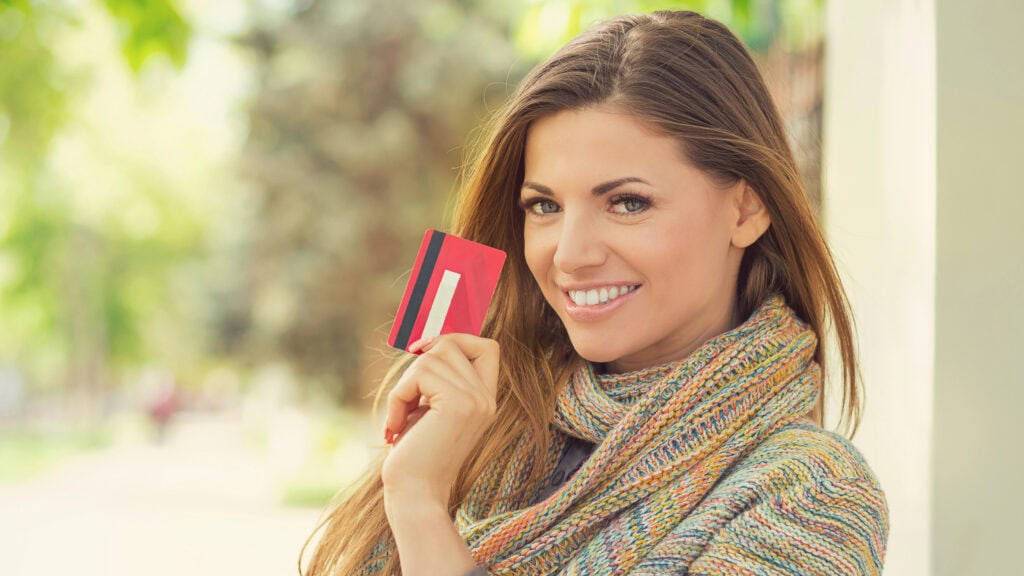 A person smiling and holding a credit card outdoors, wearing a multicolored sweater.