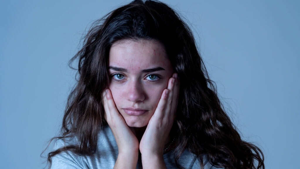 A person with long, wavy hair rests their face in their hands, looking directly at the camera with a neutral expression. They are wearing a gray shirt against a plain blue background.