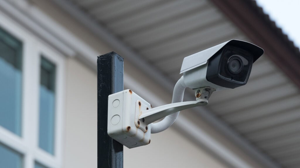 Outdoor surveillance camera mounted on a metal pole, positioned near a building with visible windows and roof.