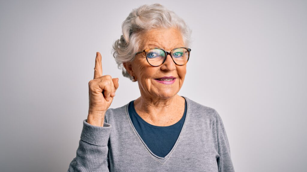 Elderly woman with grey hair and glasses, wearing a grey sweater, is raising her right index finger, smiling with a thoughtful expression, against a plain background.