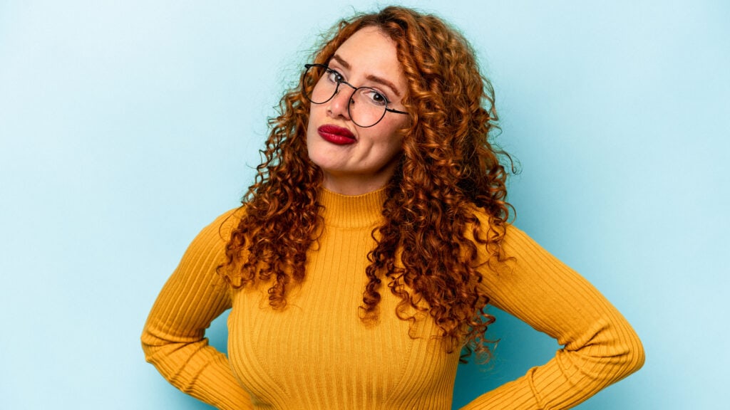 A person with curly red hair and glasses wears a mustard yellow sweater and stands against a light blue background with hands on hips.