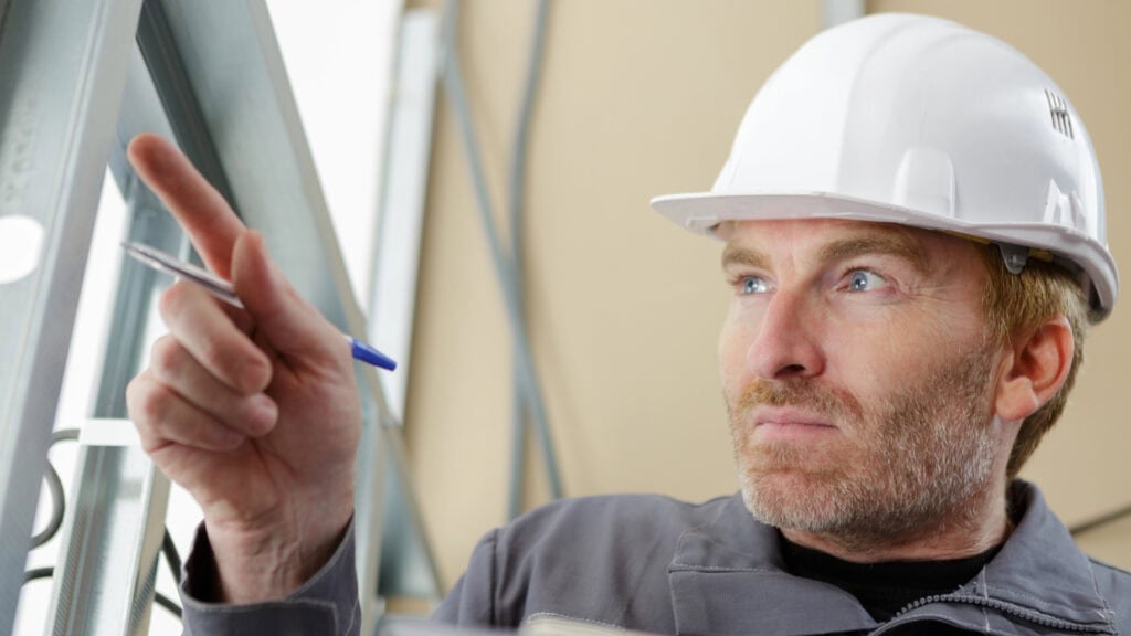 A man in a hard hat points while holding a clipboard and pen, appearing to inspect something.