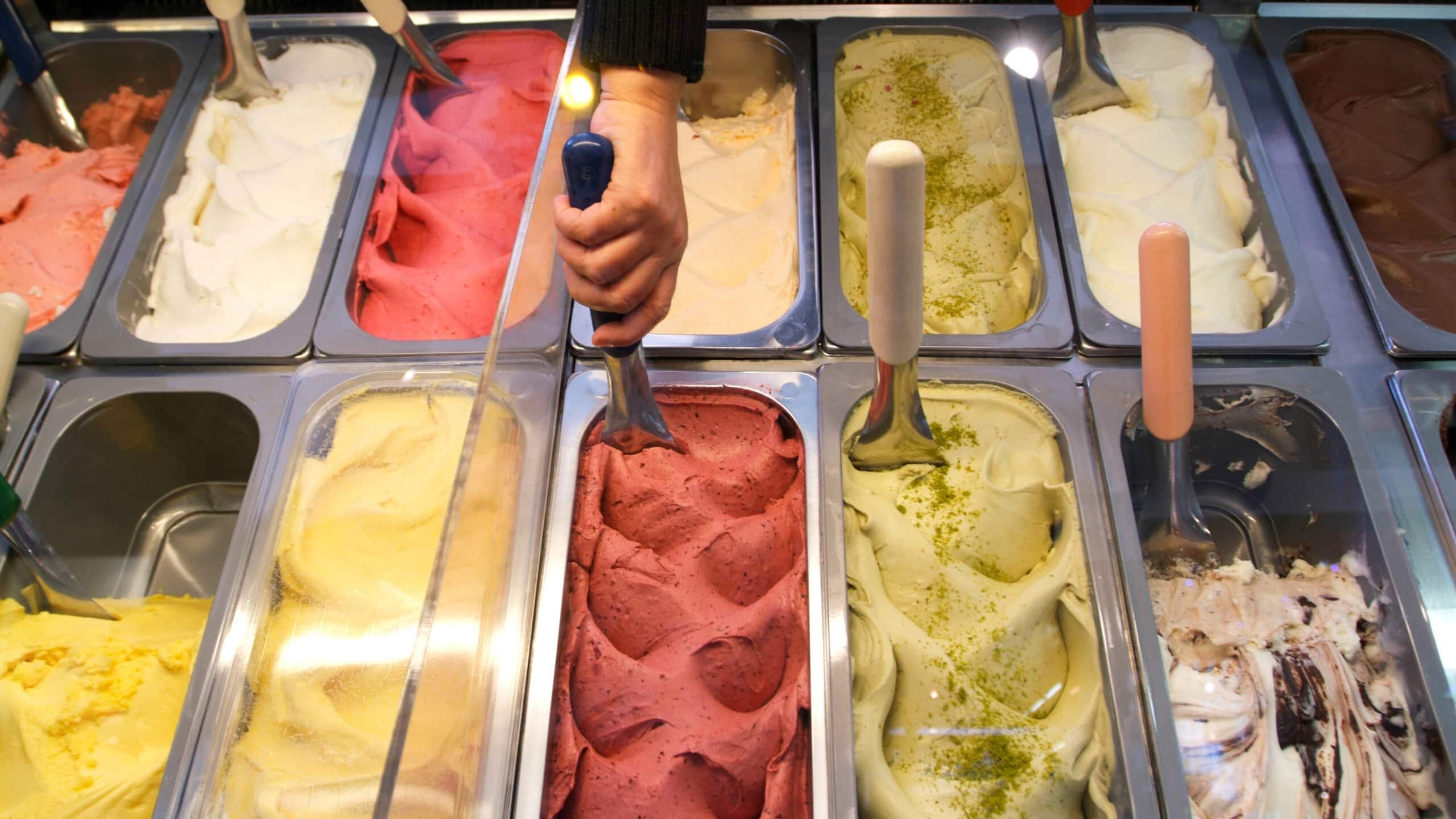 A person scoops red gelato from a display containing various flavors, including yellow, pink, white, green, and swirled options.