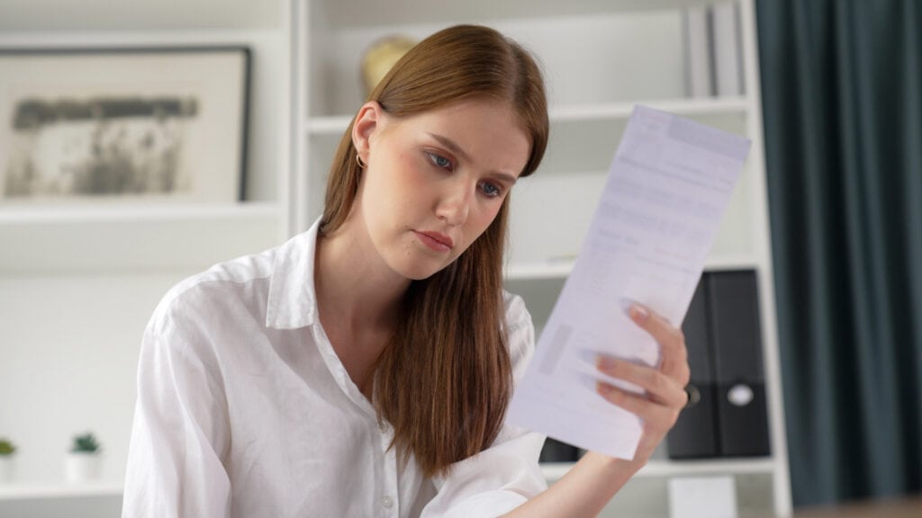 A woman in a white shirt reads a document, sitting in a room with white shelves and a framed picture in the background.