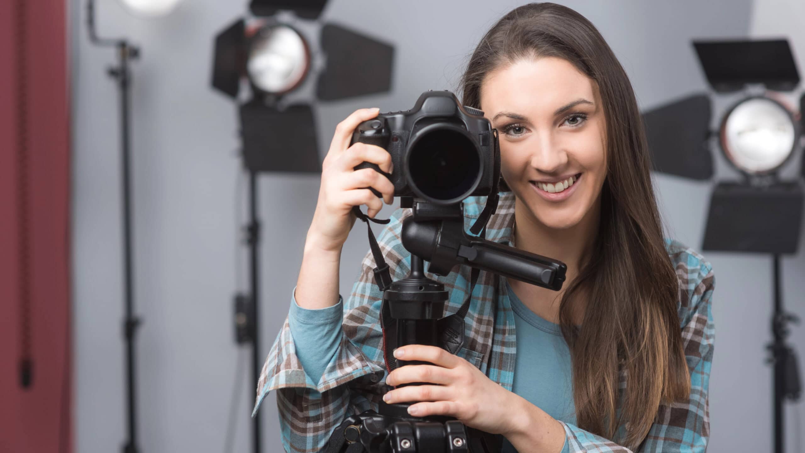 A woman smiling, holding a camera on a tripod in a photography studio with lighting equipment in the background.