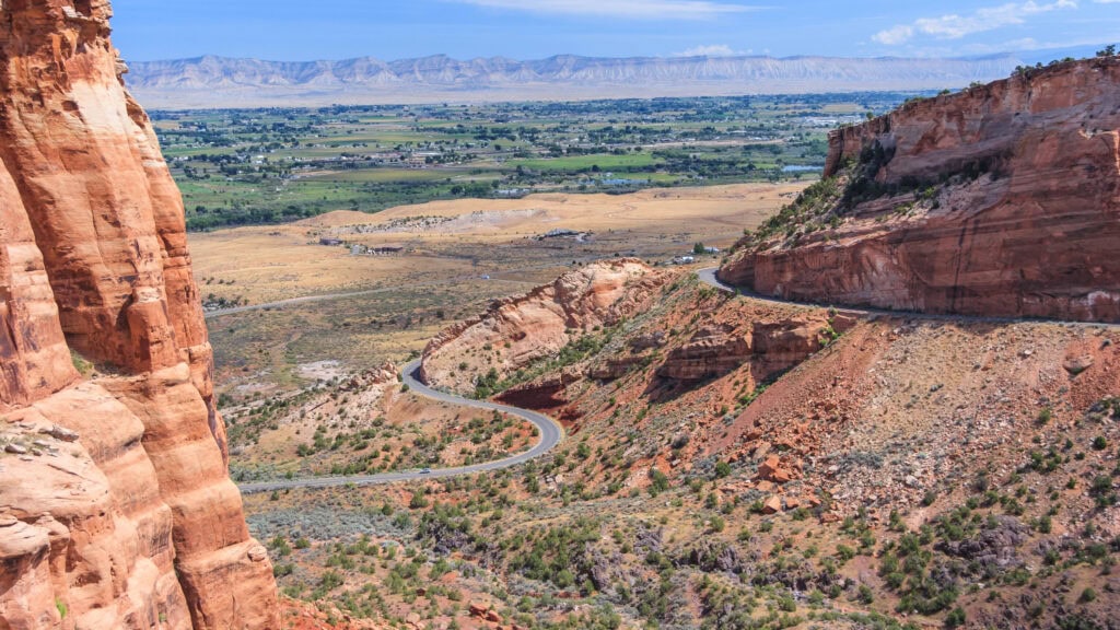 A winding road cuts through a rocky canyon landscape, with expansive flatlands and distant mountains under a partly cloudy sky.