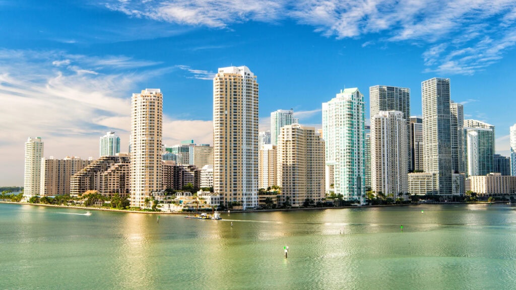 Skyline of a city with tall skyscrapers reflecting in calm water under a blue sky with scattered clouds.