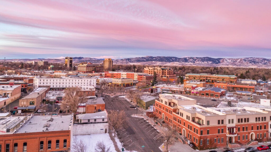 Aerial view of a small city at sunset, featuring various buildings, snow-covered streets, and distant mountains under a pink and purple sky.