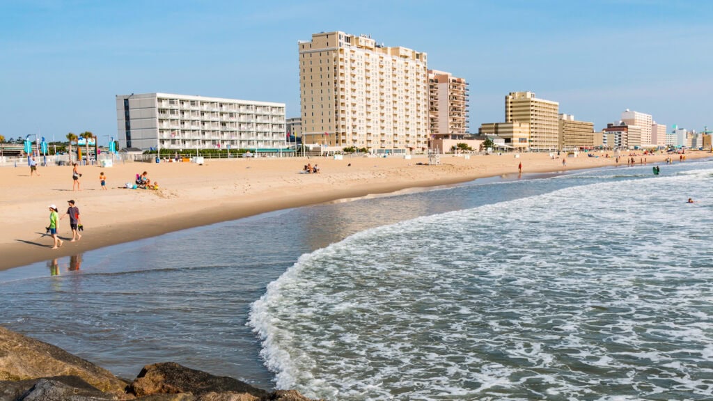 Sandy beach with gentle waves, people walking, and tall buildings in the background under a clear sky.