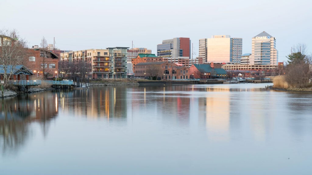 A calm river reflects surrounding modern and brick buildings in an urban landscape under a clear sky.