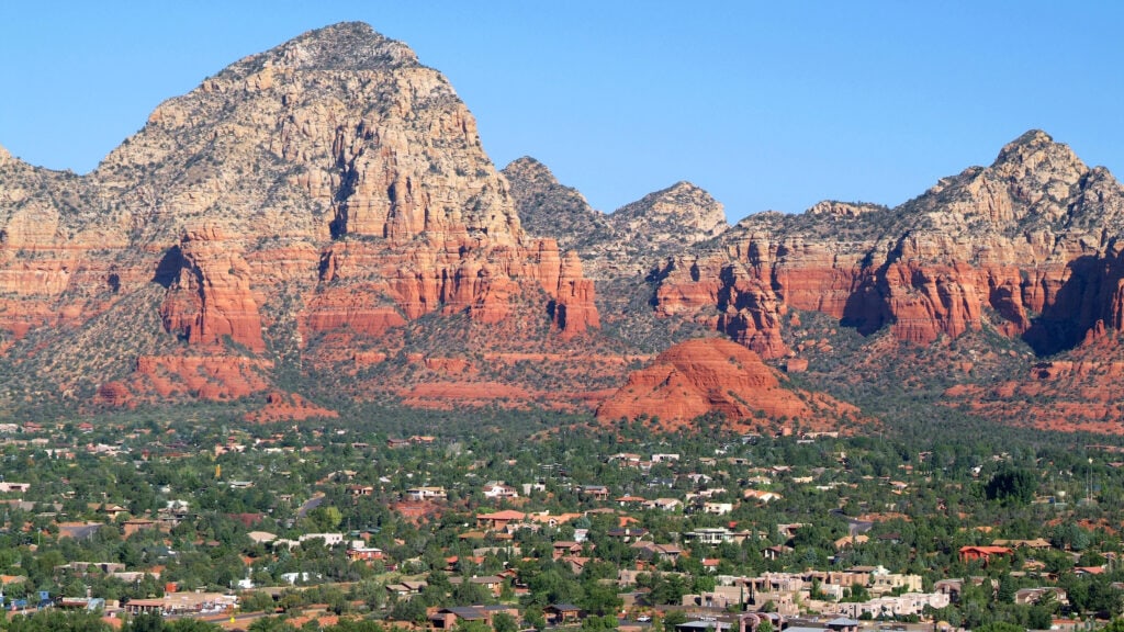 A residential area with scattered houses and greenery sits in the foreground of a rugged, red rock mountain landscape under a clear blue sky.