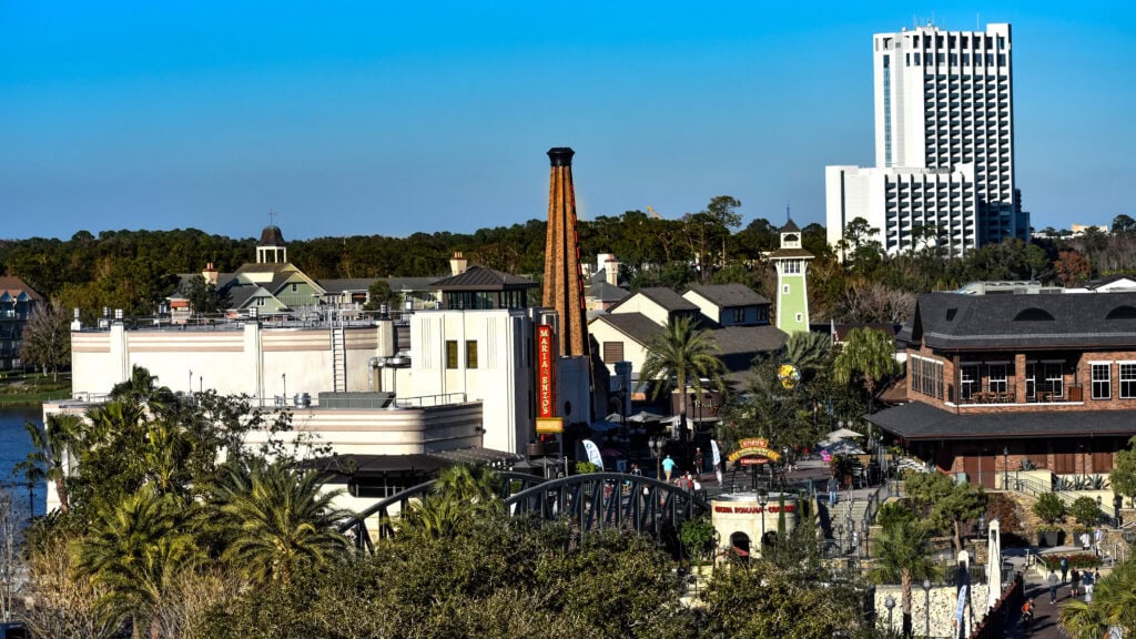 A view of an urban area with greenery, a prominent smoke stack, buildings, and a tall white hotel or apartment building in the background under a clear blue sky.