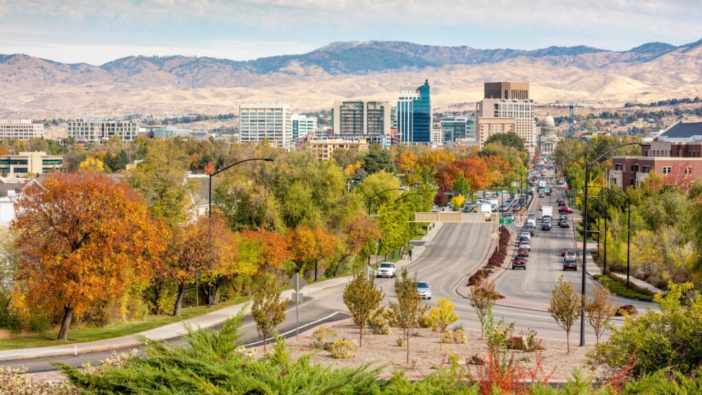 Cityscape with autumn trees, buildings, and distant hills under a cloudy sky. Busy road runs through the center, lined with foliage.