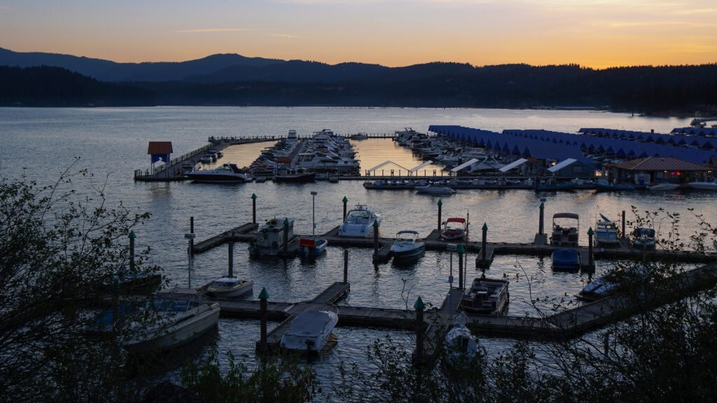 Boats docked at a marina on a calm lake during sunset, with silhouetted hills in the background and gentle ripples in the water.