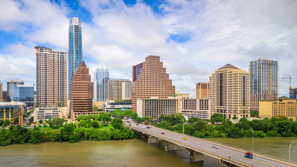 City skyline with modern skyscrapers under a partly cloudy sky, a river in the foreground with a bridge, and cars crossing.
