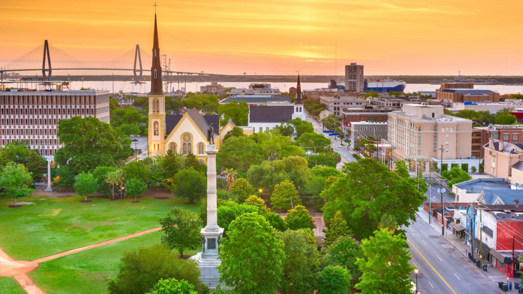 Aerial view of a cityscape with a church, green trees, and buildings at sunset. A bridge spans the water in the background.