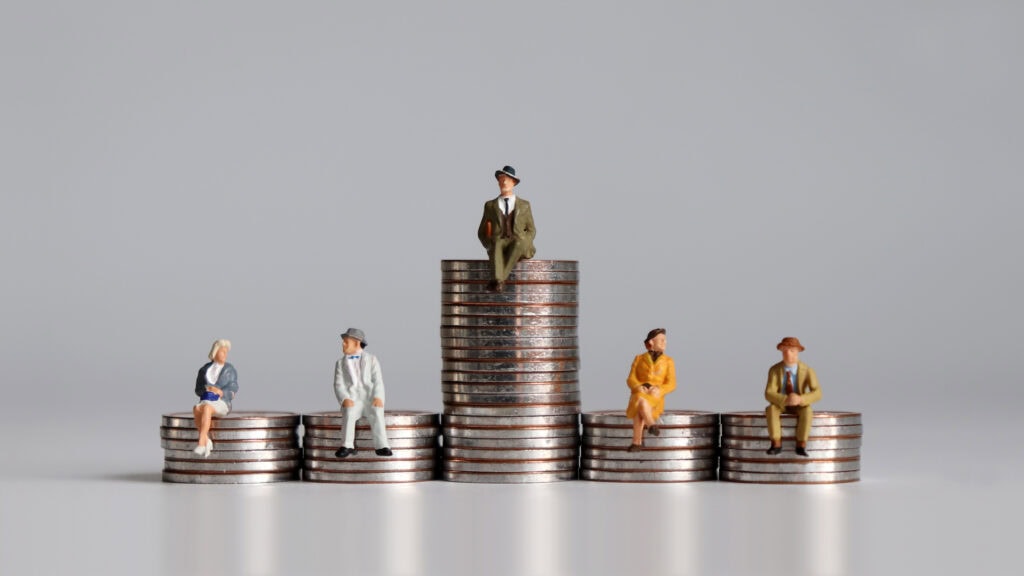 Miniature figures in business attire sit atop stacks of coins of varying heights against a plain gray background.