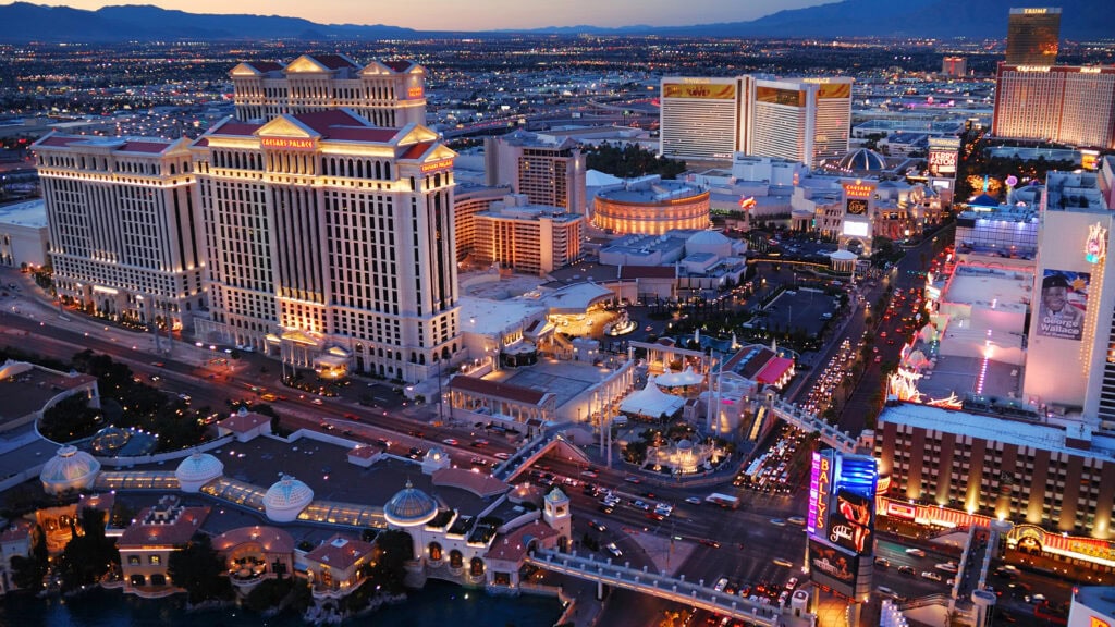 Aerial view of Las Vegas Strip at dusk, featuring illuminated hotels and casinos, including a large building with columns and domes. City lights are visible in the background.