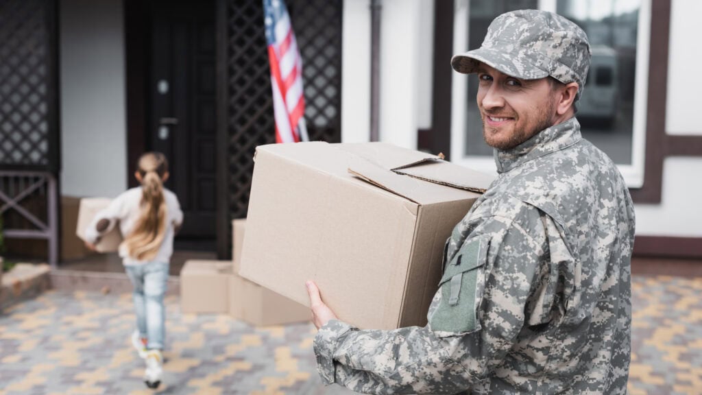 A person in military uniform carries a box outside a house with an American flag. A child carries another box in the background.