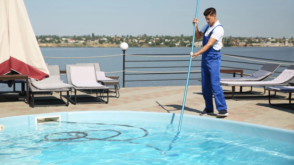 Person wearing blue overalls cleaning a swimming pool with a long pole net. Lounge chairs are nearby, with a view of a large body of water in the background.