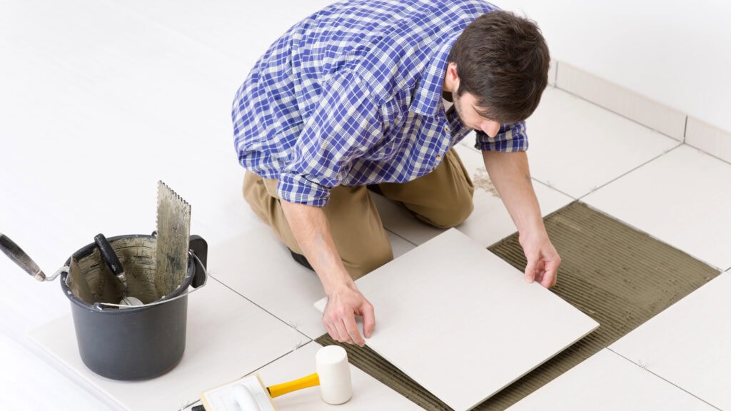 Person in a plaid shirt kneeling and laying tiles on a floor. A bucket with tools sits nearby.
