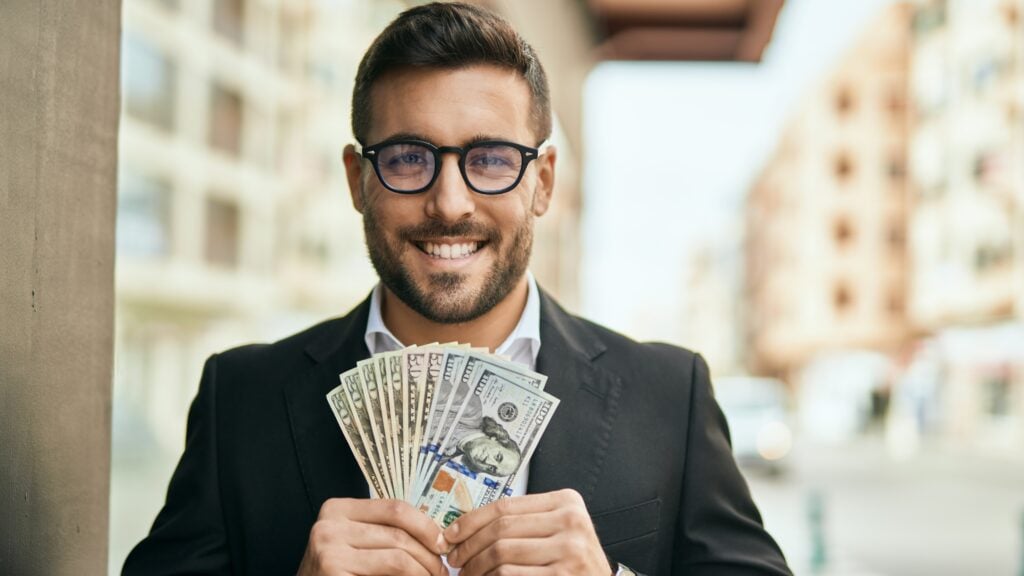 A man in a suit and glasses holds a fan of U.S. hundred-dollar bills while standing on a city street.