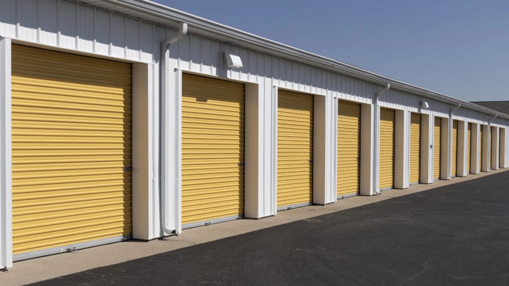 A row of storage units with closed yellow garage doors, set against a white building, beneath a clear blue sky.