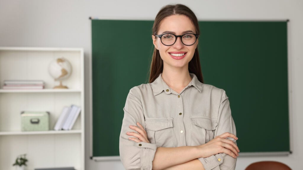 A person with glasses stands smiling with arms crossed in a classroom. A chalkboard is in the background, and shelves with books and a globe are on the left.