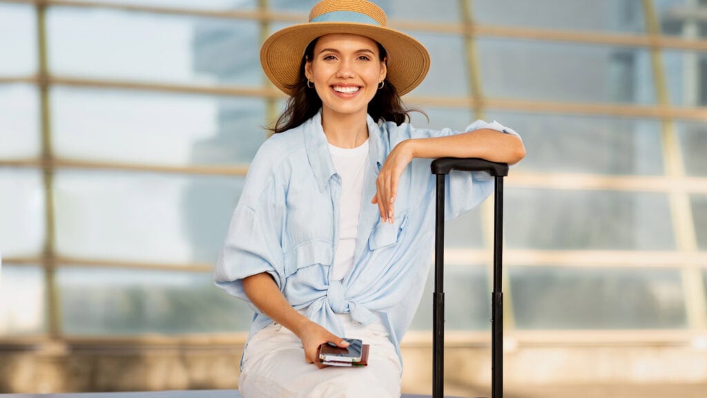 A woman in a straw hat and casual attire sits on a bench with a yellow suitcase and holds a phone and passport. She is smiling and appears relaxed.