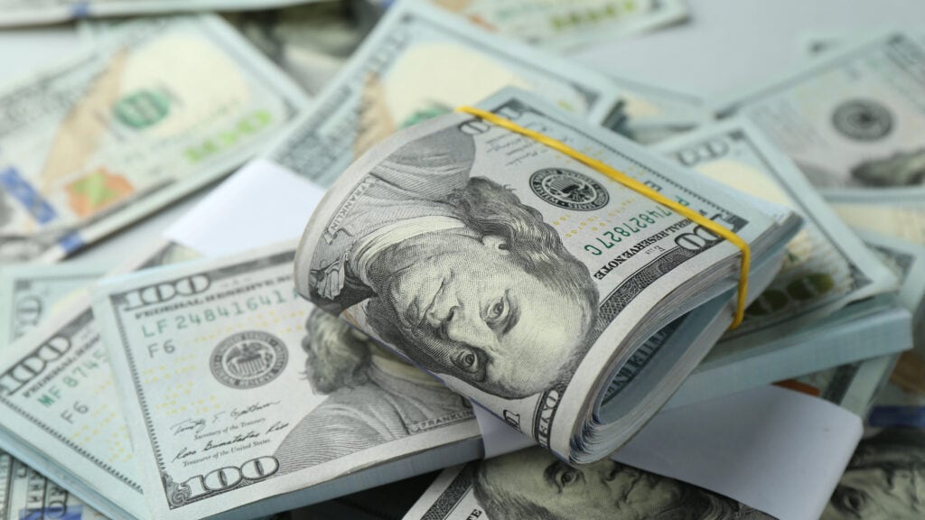 A bundle of U.S. hundred-dollar bills secured with a rubber band is placed on top of scattered bills.