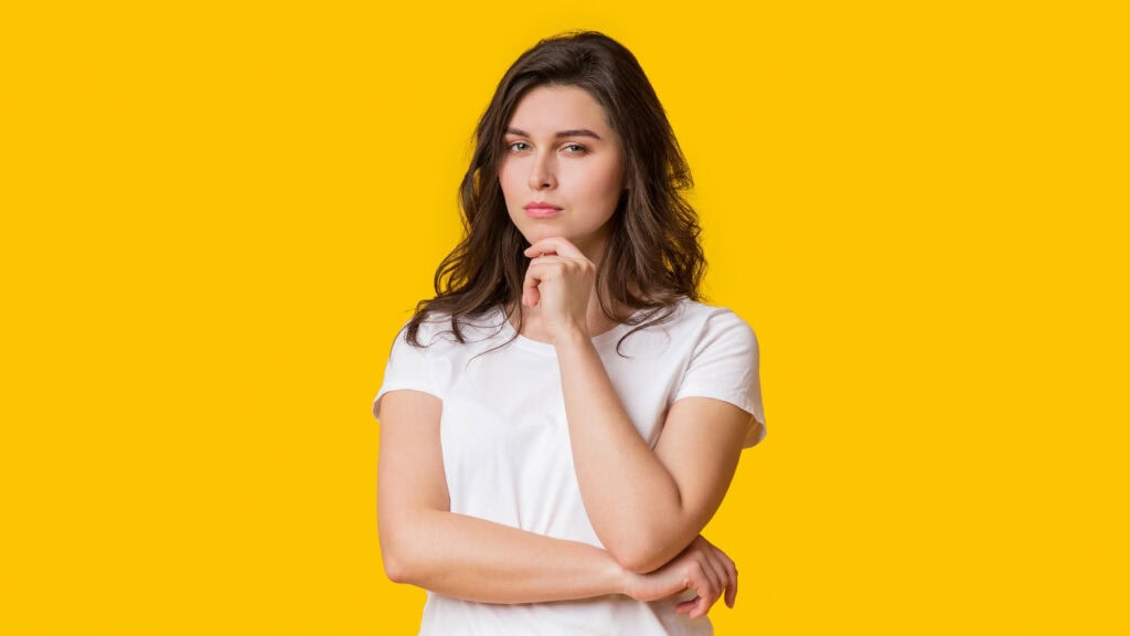 Person in a white shirt standing against a yellow background with a thoughtful expression, resting their chin on their hand.