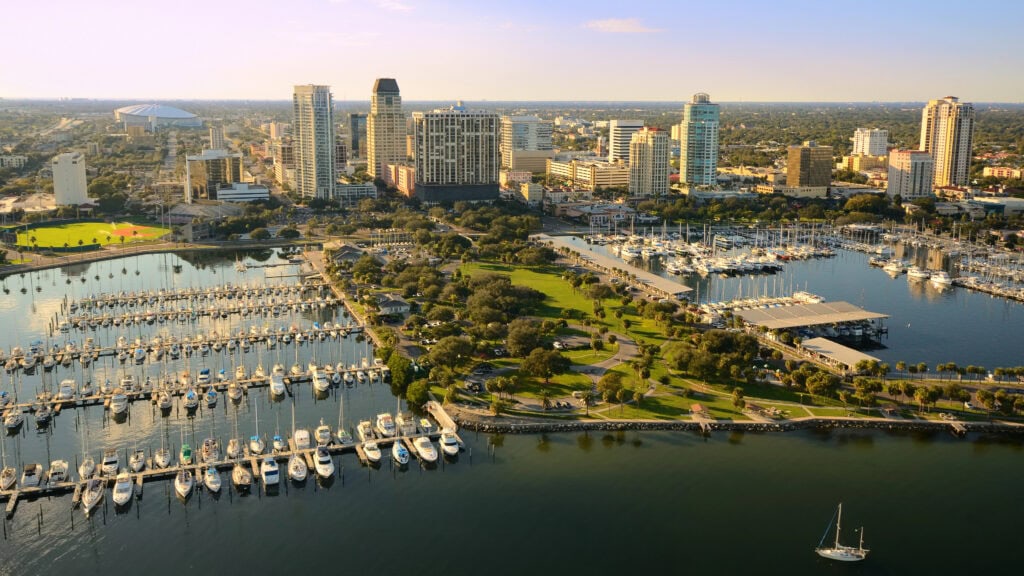 Aerial view of a coastal city with a marina filled with boats in the foreground, high-rise buildings in the center, and a stadium visible on the left.