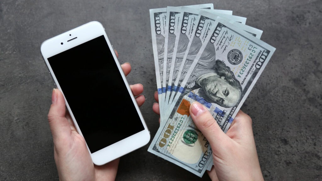 A person holds a smartphone in one hand and several U.S. hundred-dollar bills in the other hand against a gray background.