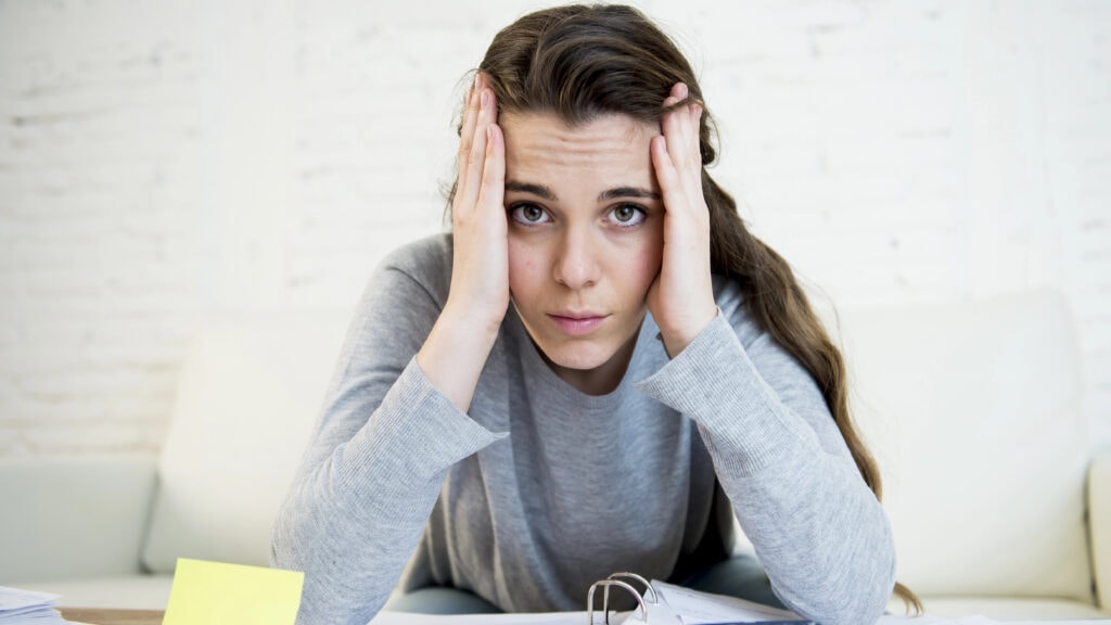 A woman sits at a table with papers, a binder, sticky notes, and a calculator, holding her head with both hands, looking stressed.