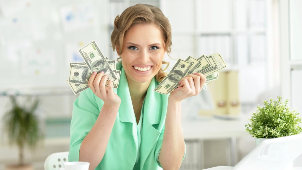 A woman in a green blazer smiles while holding stacks of US dollar bills in both hands in an office setting.