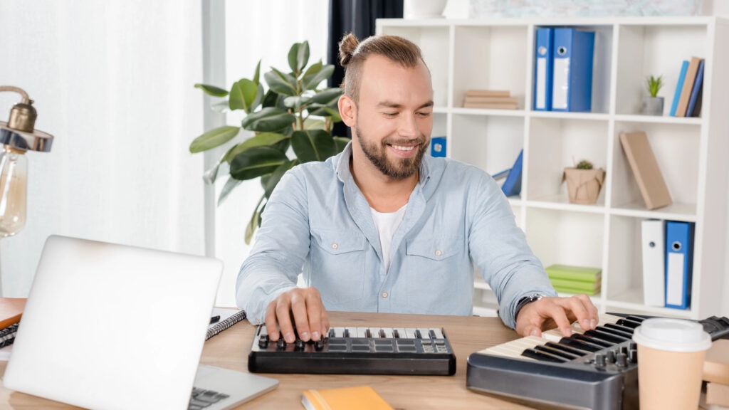 A man with a bun uses a keyboard and a MIDI controller at a wooden desk, surrounded by a laptop, coffee cup, and books.
