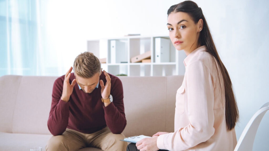 A woman holding a clipboard looks at the camera while a man in a maroon sweater sits on a couch with his head in his hands, appearing stressed.