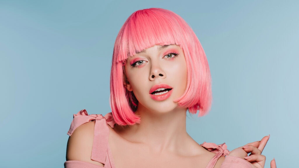 Person with pink hair and pink eye makeup poses against a blue background, wearing a pink top with ribbon sleeves.
