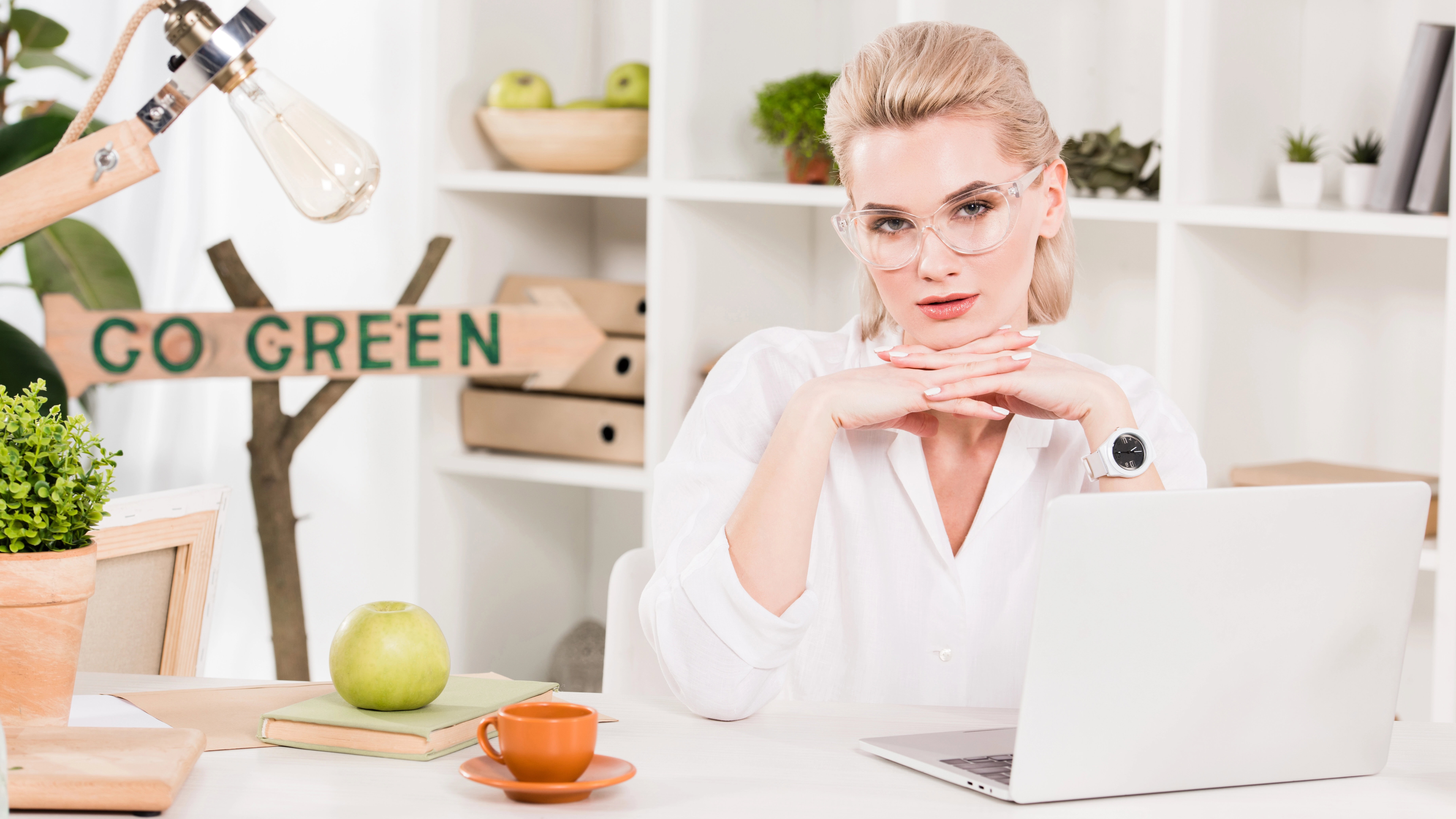 A person in glasses sits at a desk with a laptop, books, and a green apple. A "GO GREEN" sign and potted plants are in the background.