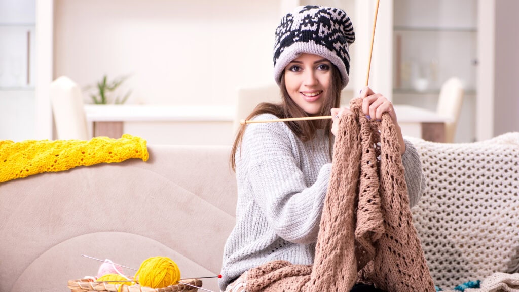 A person wearing a knitted hat is sitting on a sofa, knitting a brown blanket. A basket with yellow and pink yarn is on the sofa.