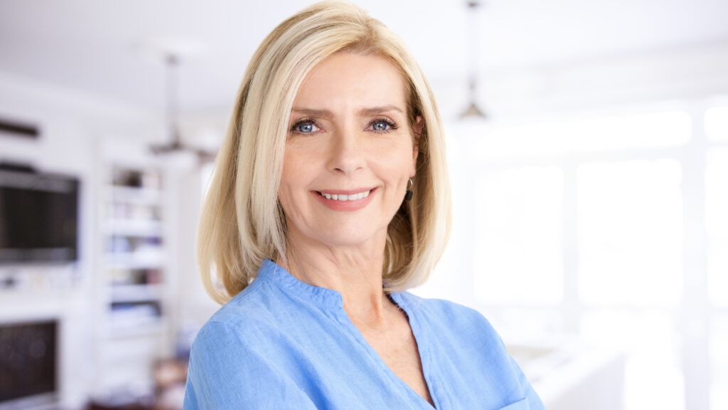 A woman with blonde hair and a blue shirt smiles in a bright room.