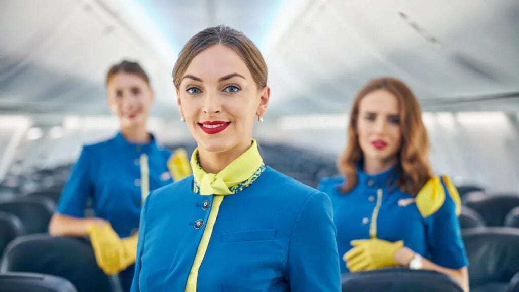 Three flight attendants stand in an airplane aisle, wearing blue uniforms with yellow accents. The focus is on the attendant in the foreground, smiling at the camera.