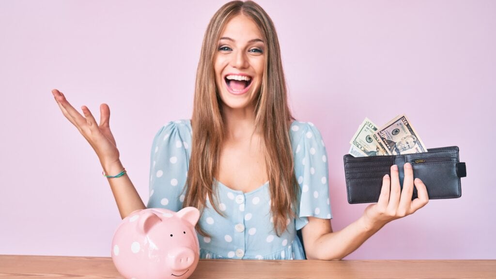 Young blonde girl holding wallet with dollars and piggy bank