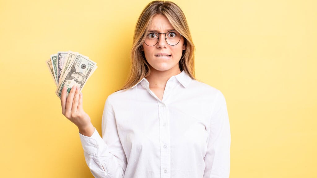 A person with long hair and glasses holds a fanned-out stack of U.S. dollar bills, looking concerned against a yellow background.
