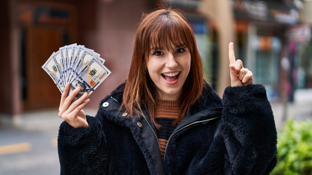 A woman with brown hair is smiling and holding a fan of US $100 bills in one hand while pointing upwards with the other hand. She is wearing a black coat and standing outdoors.