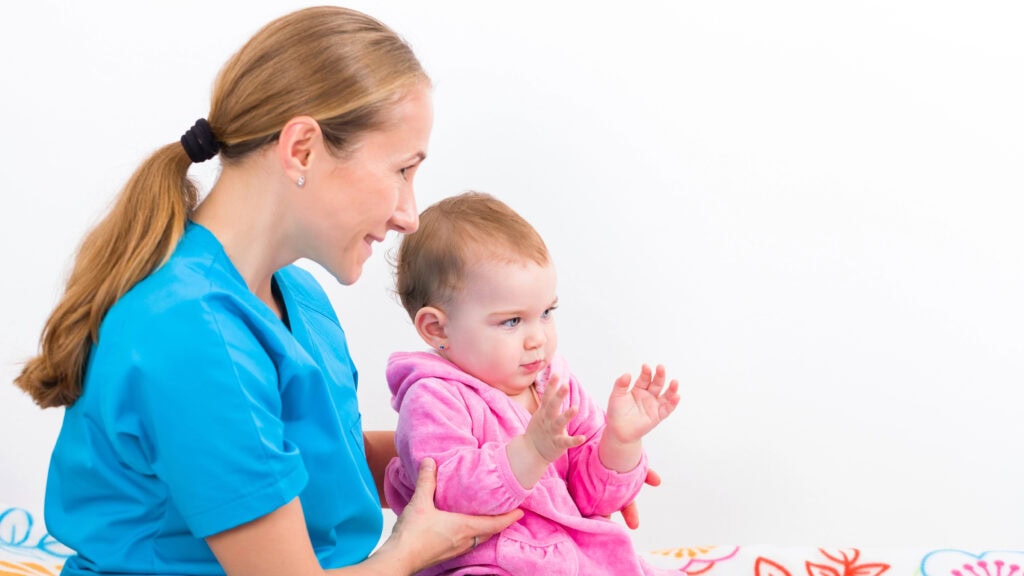 A woman in blue scrubs sits with a baby in pink clothes on a colorful blanket, guiding the baby’s hands.