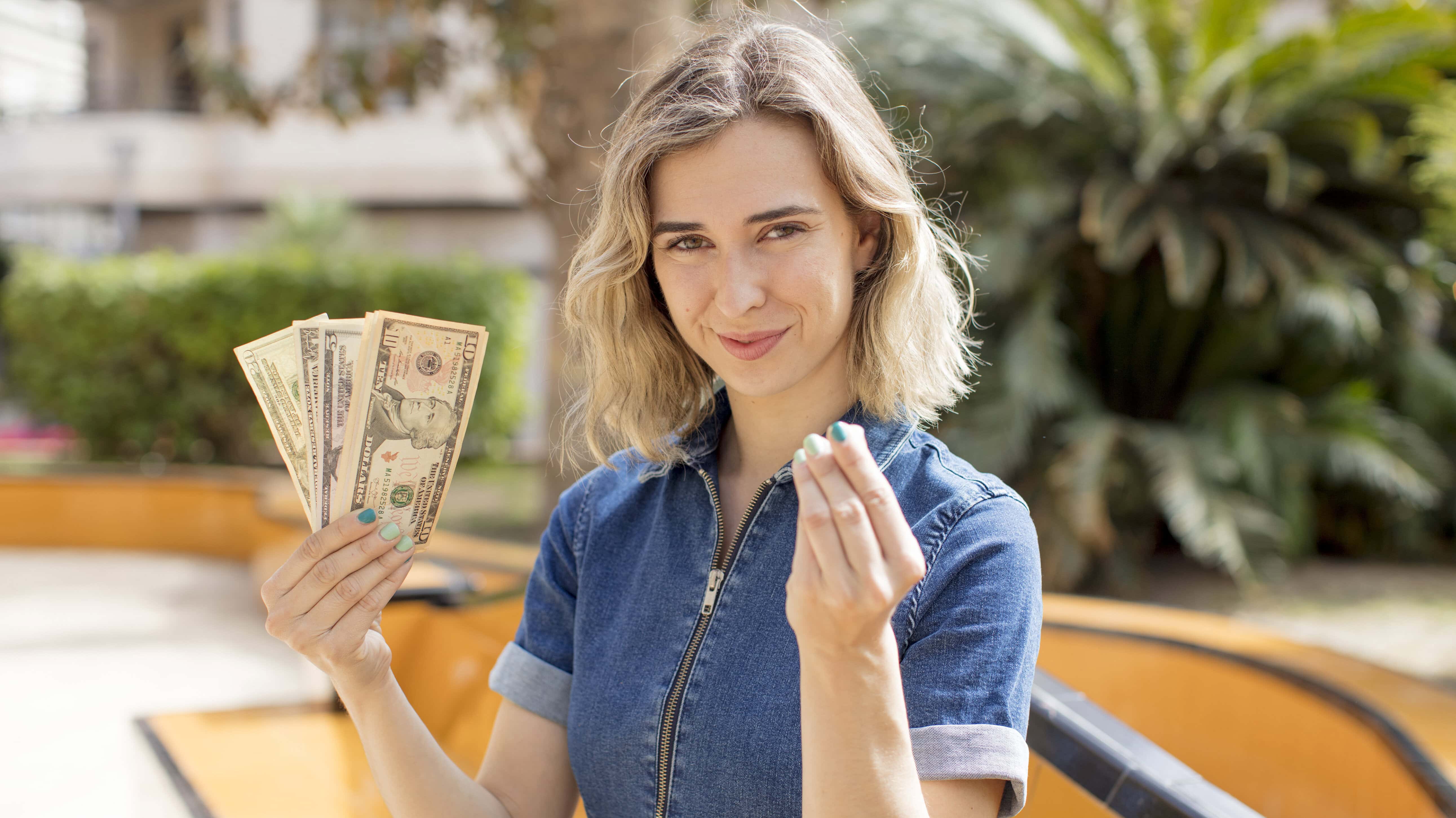A woman in a denim outfit holds several U.S. dollar bills and gestures with her other hand, outdoors near greenery.