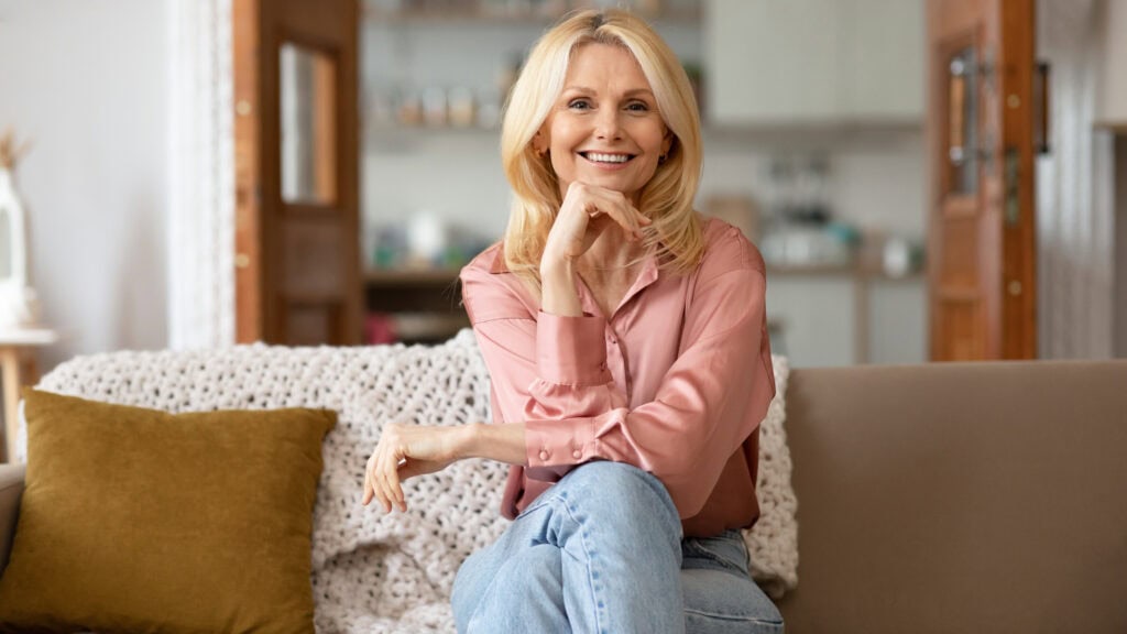 A woman with blonde hair sits on a sofa, wearing a pink blouse and blue jeans, smiling with her hand under her chin. The background features a cozy indoor setting.