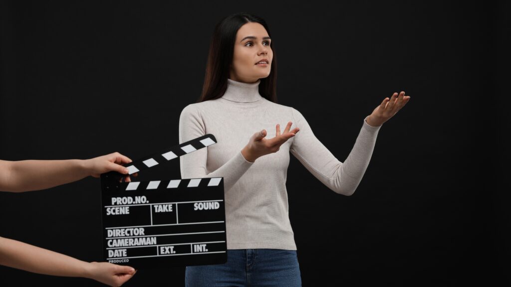 A woman in a light turtleneck holds her arms out, standing in front of a clapperboard against a black background.